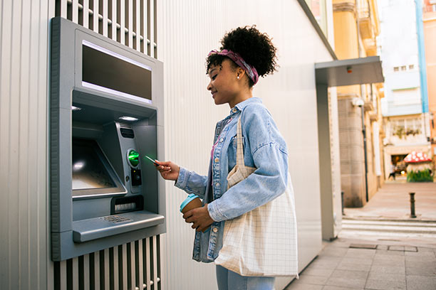 A young Black woman carries a tote bag and uses an ATM.