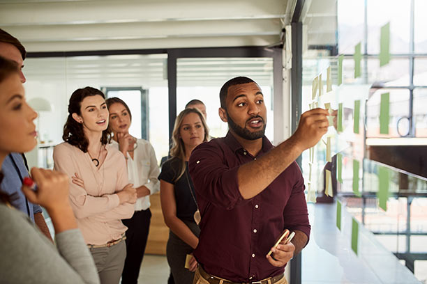 A Black man stands with his 6 co-workers and moves sticky notes on the wall.