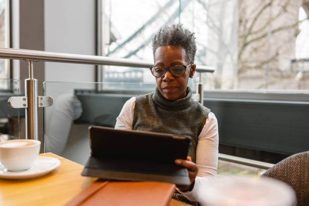 An older Black woman with short gray hair reviews her tablet while sitting at a cafe.