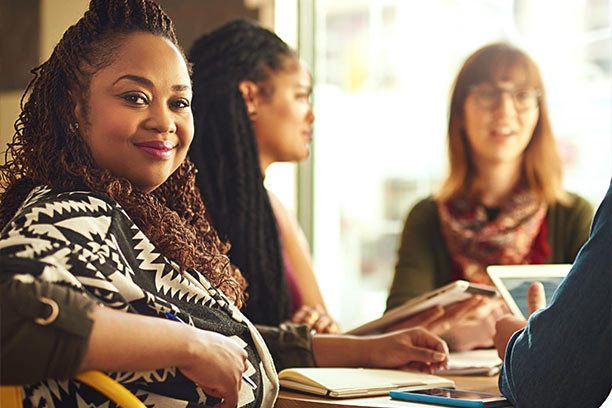 A smiling Black woman with dark curly hair smiles at the camera as two other woman discuss something behind her.