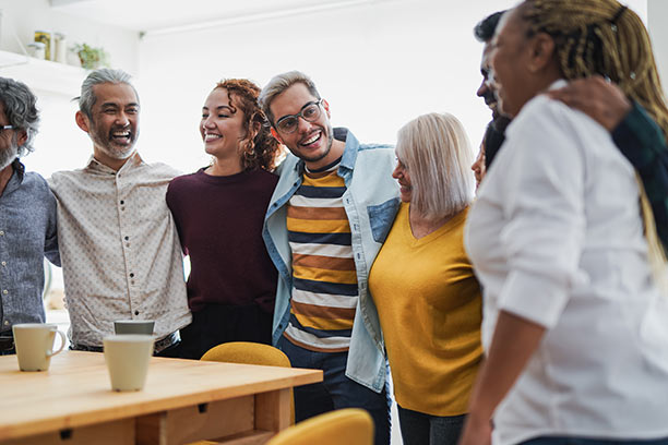 A diverse group of young and older adults stand in a semi-circle with arms over each other's shoulders.