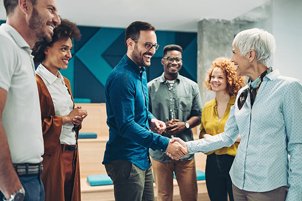 A diverse group of men and women stand together, with a young man and older woman shake hands.