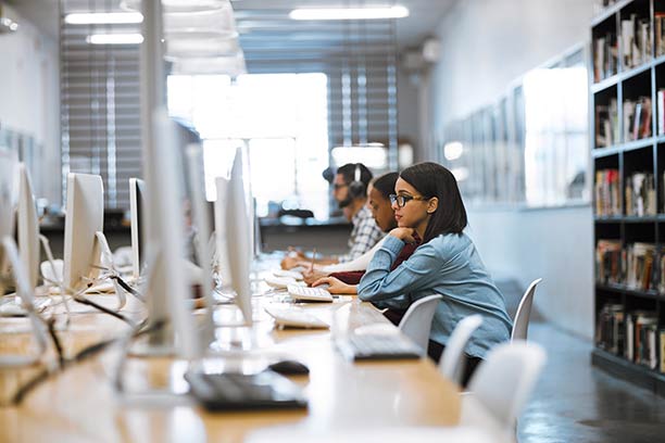 3 young adults sit at a long table of iMacs, working on the computers.