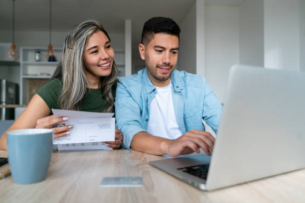 A young woman and man sit at a table and review their joint finances on their laptop screen.