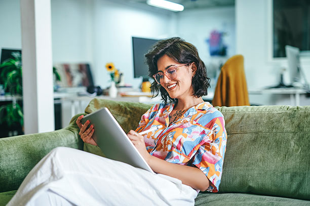 A young woman with dark hair and light brown skin wearing a colorful top sits on a green couch and reviews her tablet.