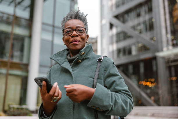 An older Black woman with short gray hair walks outside in a city holding her cell phone.