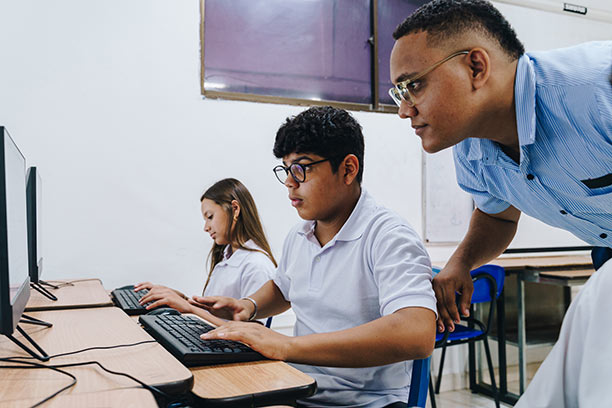 A young boy with light brown skin sits next to a classmate at a computer lab while his male teacher leans over to help him.