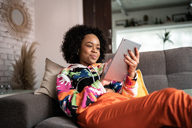 A young Black woman with dark curly hair sits on her couch while looking at her tablet.