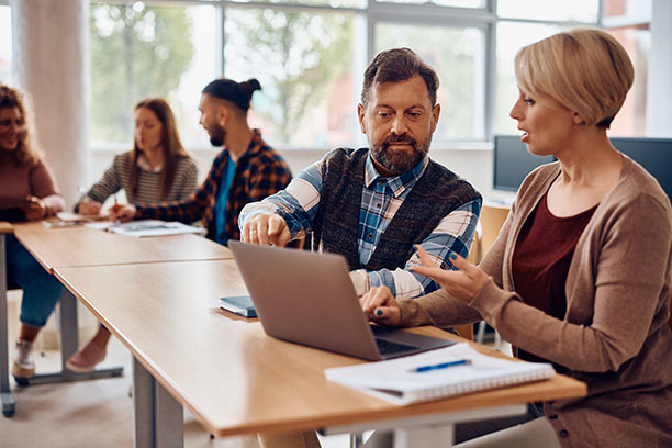 An older white man and white woman review something on a laptop screen in a conference room setting.