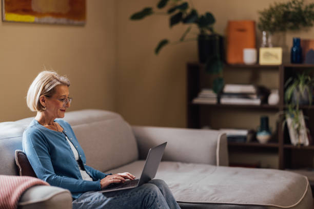 An older white woman sits on a beige couch and reviews something on her laptop.