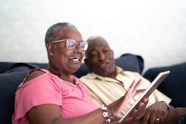 A Black man and woman sit together on the couch, smiling and looking at something on an iPad.