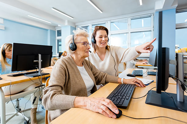 An older white woman sits at a computer in a computer lab while a young woman instructor helps her.