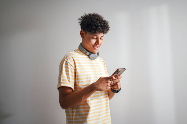 A young man with dark hair and light brown skin looks down at his cell phone with headphones hung around his neck.