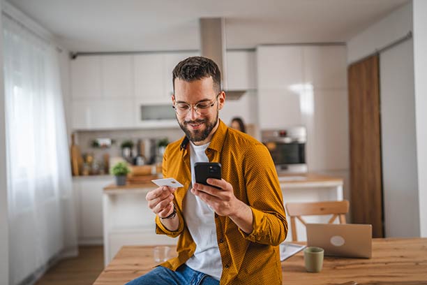 A white man stands in his kitchen, smiling at his cell phone.