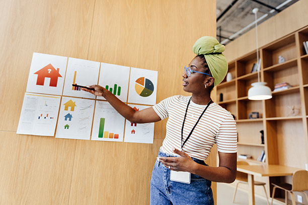 A young Black woman wearing a light green hair wrap points to a series of 8 infographics taped to the wall.