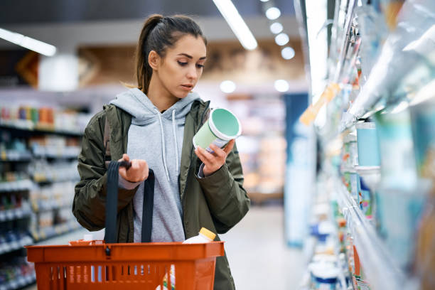 A woman holds a shopping basket and reads a nutrition label. 