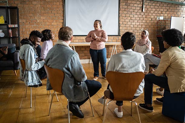 A young woman stands at the center of a semi-circle of other young people sitting in chairs.