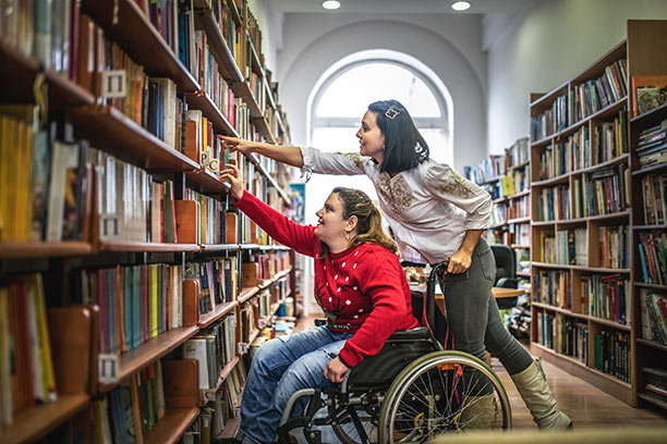 A white woman in a wheelchair reaches for a book on a library shelf, with another woman reaching for the shelf as well from behind the wheelchair.