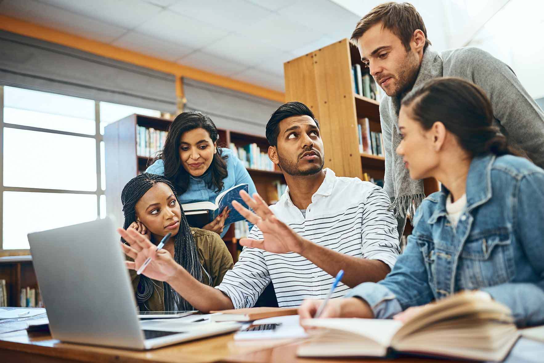 A diverse group of 5 adults sit together in a library and discuss various materials on the table.