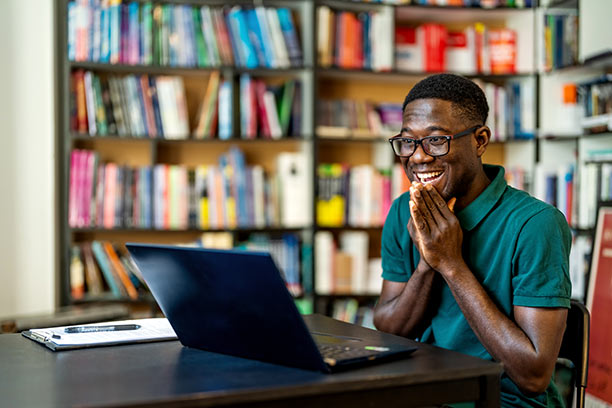 A young Black man sits at a table in front of a large bookcase, smiling at something he sees on his laptop with his hands excitedly brought up to his face.