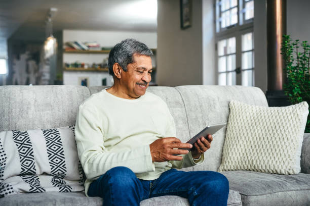 An older man with light brown skin and gray hair sits on a couch and looks at a tablet.