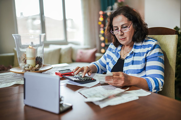 A woman with dark hair and glasses sits at a table reviewing documents and using a calculator.