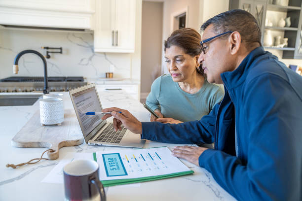 A husband and wife review a laptop screen between them while they sit at a kitchen island.