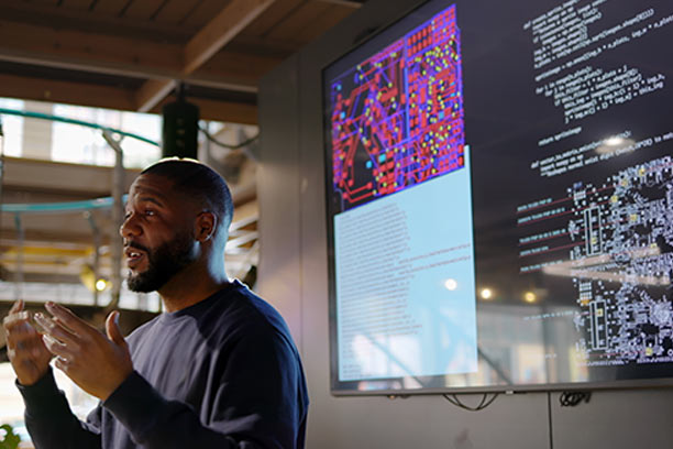 A young Black man stands in front of a projected presentation and gives a lecture.