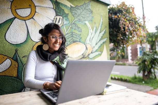 A young woman with dark brown skin and dark hair smiles outside in front of a mural while working on a laptop.