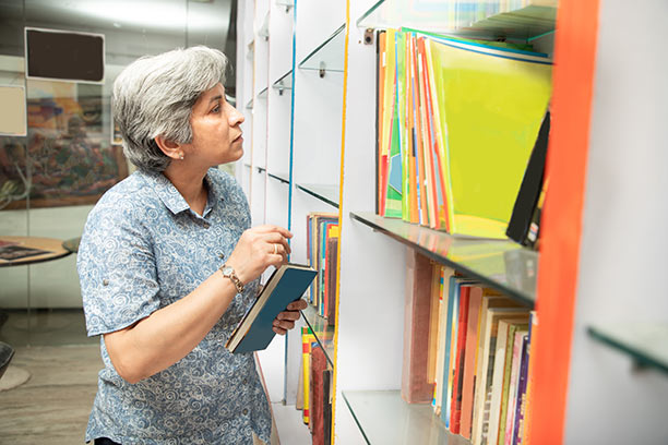 An older woman with short gray hair and light brown skin reviews files that are organized on a shelf. 