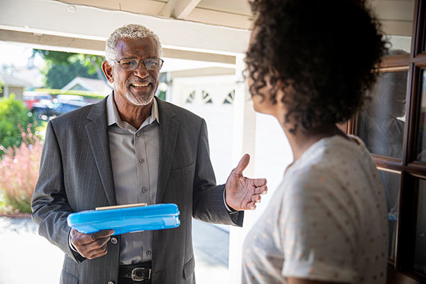 An older Black man with gray hair, glasses, and wearing a suit jacket talks to someone while holding a blue clipboard.