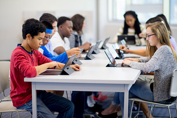 A diverse group of young students sit at a long table and work on laptops.