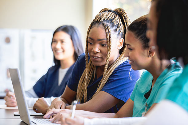 A diverse group of female students wearing blue scrubs collaborate on a laptop.