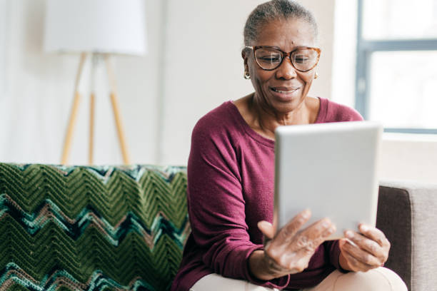 An older Black woman sits on a green couch and looks at a tablet.