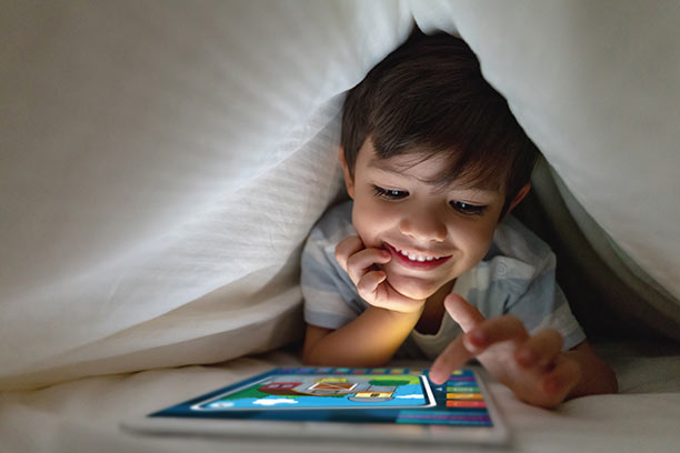 A young boy with light skin and short dark hair plays a game on his tablet in a blanket fort.