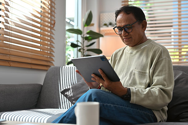 An older man with short dark hair, glasses, and light brown skin sits on a couch and reviews information on a tablet.