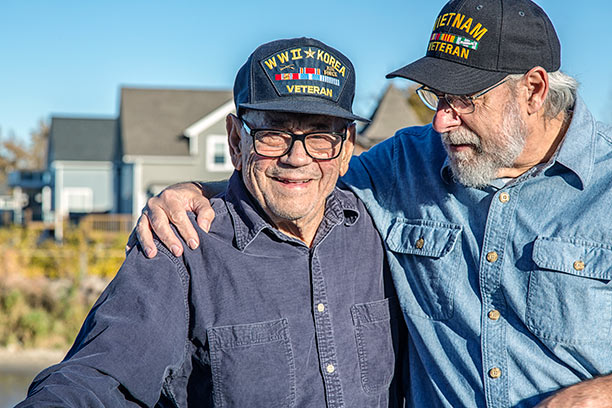 Two older white men, one with his arm over the other's shoulder, smile while wearing Veteran hats.