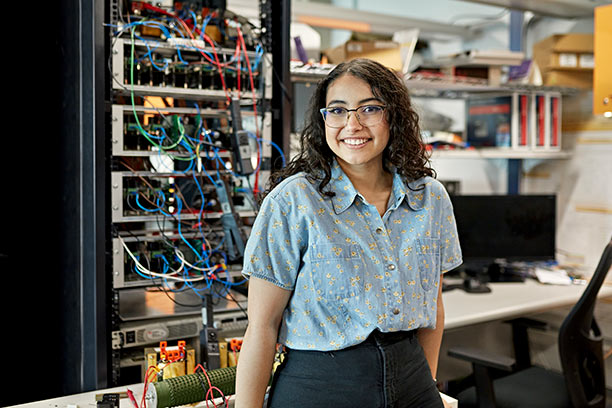 A young woman with dark curly hair and light brown skin stands in front of a large electrical panel, smiling.