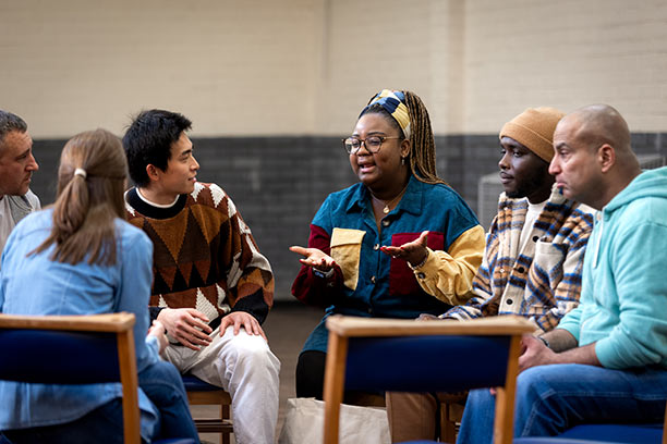 A diverse group of people sit in a circle of chairs, talking.