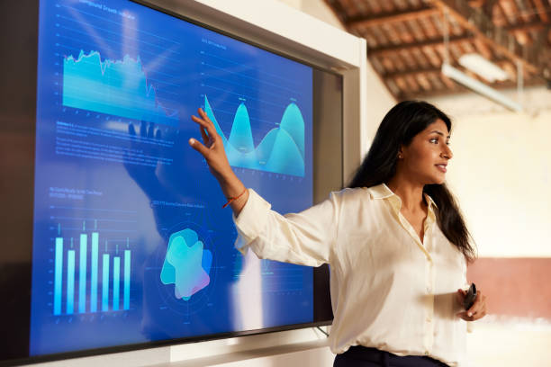 A woman with light brown skin and long, dark hair stands in front of a large digital display and presents several graphs and charts