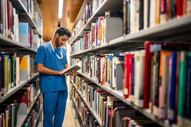 A man wearing blue scrubs stands between two library stacks of books reading a book from the shelf.