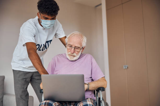 A young person with short black hair wearing a mask helps an older man with a white beard sitting in a wheelchair view something on his laptop.