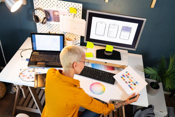 A woman with closely cropped blonde hair sits at a desk with a laptop and a larger monitor. She wears a yellow jacket and is looking at a paper report.