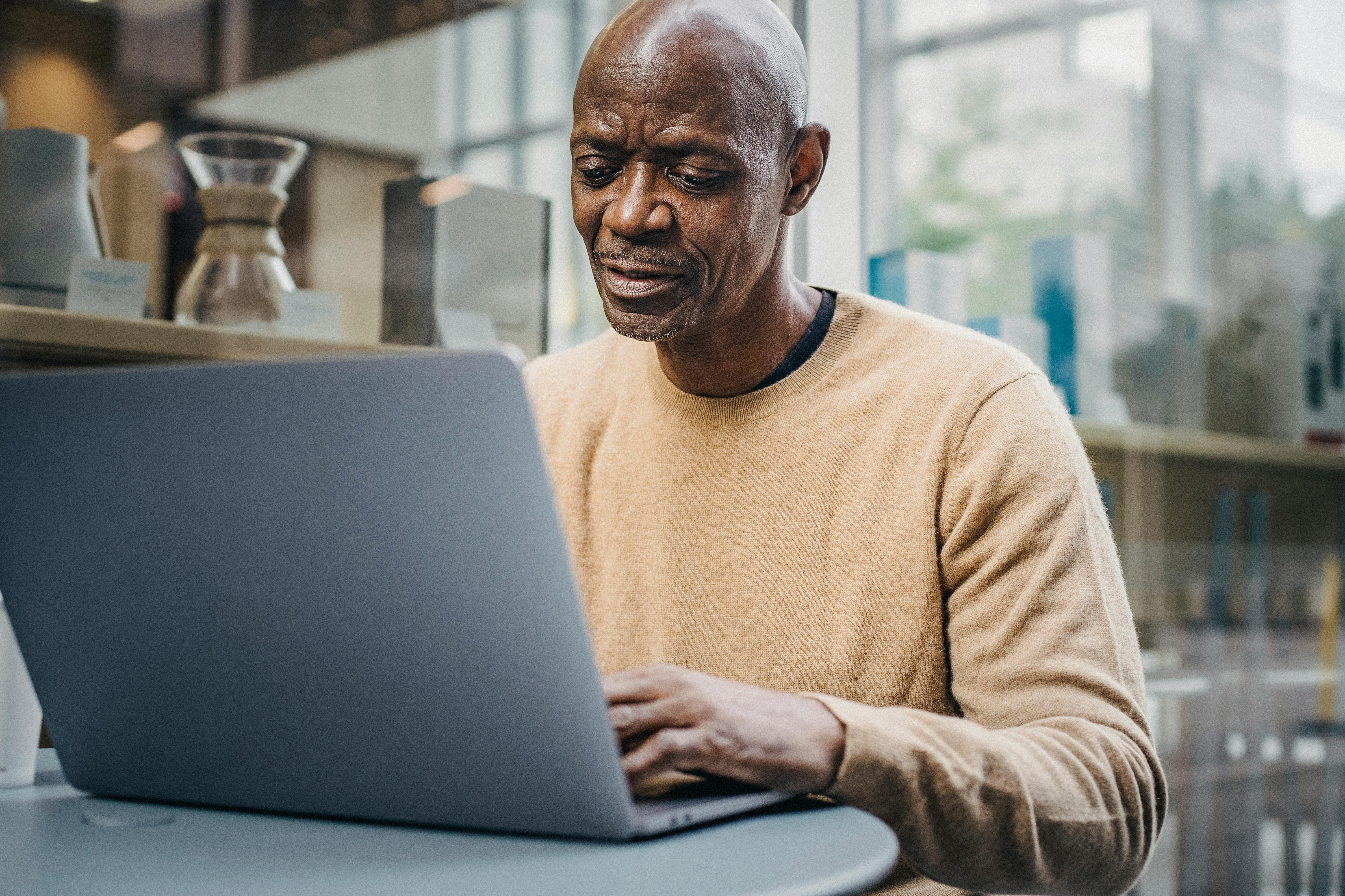 A Black man wearing a yellow sweater works on a laptop outside at a cafe table.