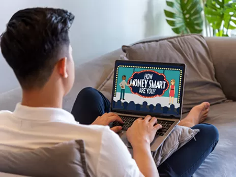 man sits on a couch using a laptop with the Money Smart image open in the browser window