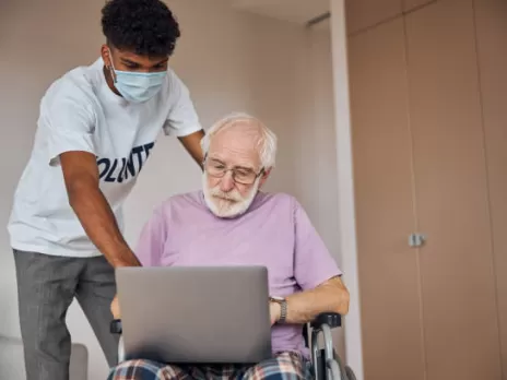 A young person with short black hair wearing a mask helps an older man with a white beard sitting in a wheelchair view something on his laptop.