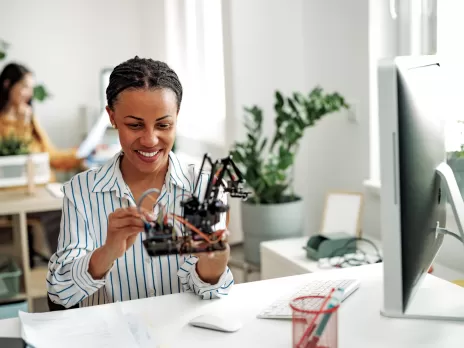 A young Black woman sits at a desk in a bright office space and works on putting together robotics parts.