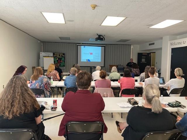 A diverse group of people sit in a conference room in three rows of tables and watch a projected presentation from PA Forward.