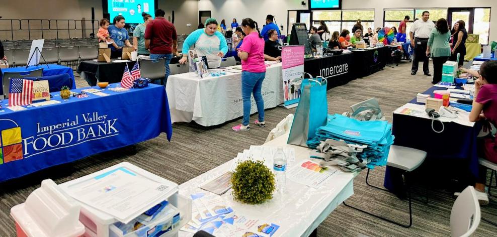 VDS en Calexico hosts a table at a community event. Community members walk around a large room, stopping by tables for various local organizations.