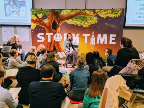 Test 1 Group of adults sitting, facing a woman reading in front of a large story time poster. 